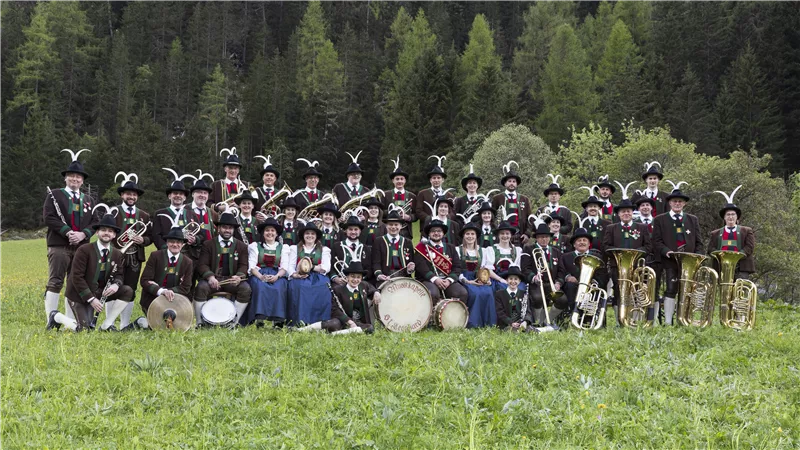 Eine Gruppe von Musikern steht auf einer Wiese, umgeben von Bäumen. Die Mitglieder tragen traditionelle Trachten und spielen verschiedene Instrumente.