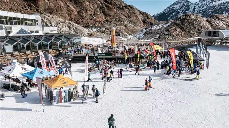Ein belebter Skihang mit vielen Menschen und bunten Zelten. Im Hintergrund sind schneebedeckte Berge und eine klare Himmel zu sehen.
