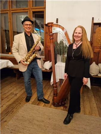 Two musicians are standing in a cozy room. The man is playing the saxophone and the woman is standing next to a harp.