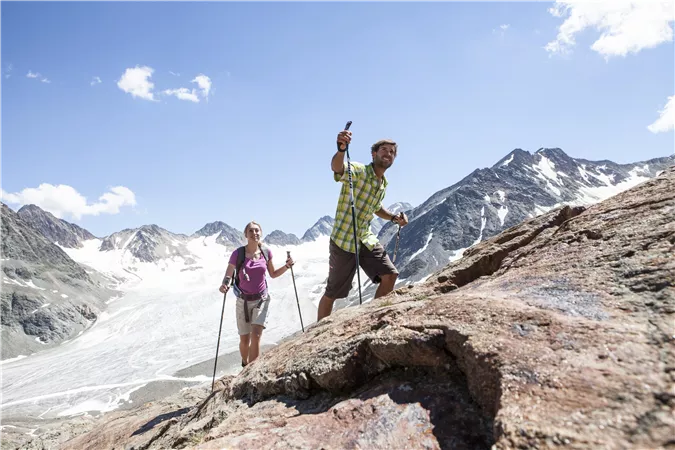 Zwei Wanderer erkunden einen Berg mit schneebedeckten Gipfeln im Hintergrund. Die Sonne scheint und der Himmel ist klar.