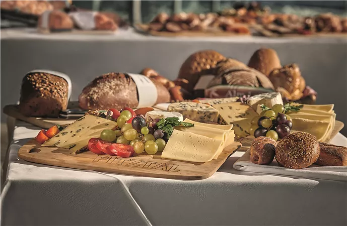 Ein schöner Tisch mit einer Auswahl an Käse, frischen Trauben und buntem Gemüse. Im Hintergrund sind verschiedene Brote und Gebäck zu sehen.