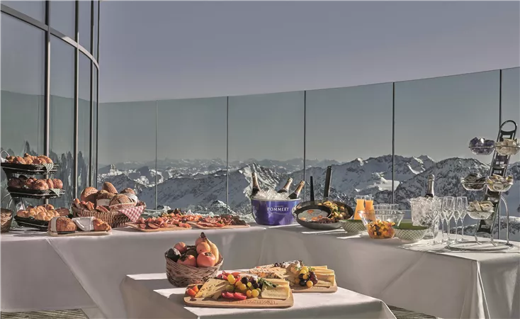 Ein festlich gedeckter Tisch mit verschiedenen Köstlichkeiten, darunter Gebäck, Obst und Getränke. Im Hintergrund sind schneebedeckte Berge und ein klarer Himmel zu sehen.