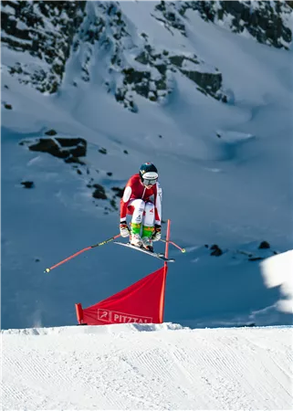 A skier jumps over a red gate in the snow. The landscape is mountainous and wintry.