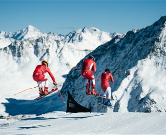Three skiers in red suits jump over a snow slope with mountains in the background. The sky is clear and blue.