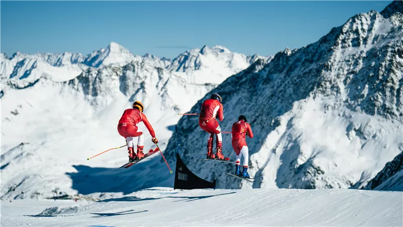 Three skiers in red suits jump over a snow slope with mountains in the background. The sky is clear and blue.
