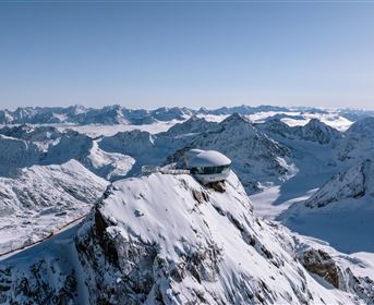 An impressive mountain landscape with snow-covered peaks and a bright blue sky. In the center stands a modern building on a high rock.