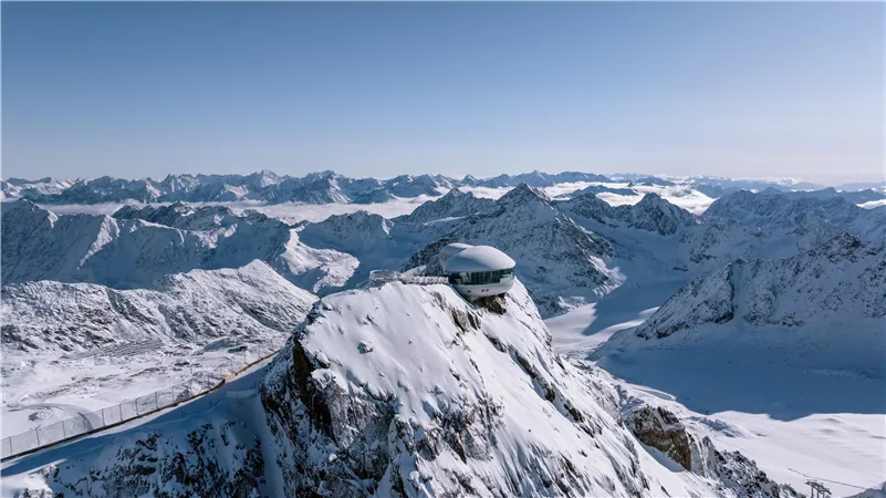 Eine beeindruckende Berglandschaft mit schneebedeckten Gipfeln und strahlend blauem Himmel. In der Mitte steht ein modernes Gebäude auf einem hohen Felsen.