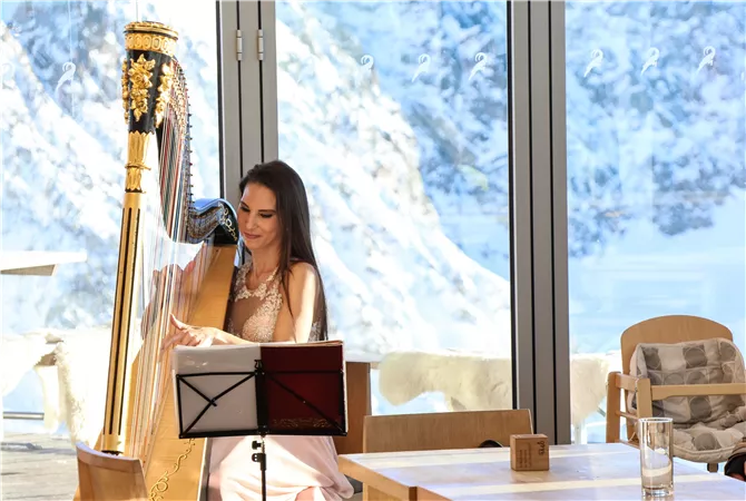 Eine elegante Frau spielt Harfe in einem modernen Raum mit großen Fenstern. Im Hintergrund sind schneebedeckte Berge zu sehen.