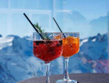 Zwei Cocktails in eleganten Gläsern, einer rot und einer orange, stehen auf einem Tisch. Im Hintergrund sind majestätische Berge und ein klarer Himmel sichtbar.