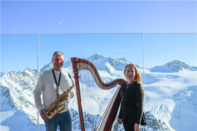Zwei Musiker stehen in den Bergen, ein Mann mit einem Saxophon und eine Frau an einer Harfe. Im Hintergrund sind schneebedeckte Gipfel und ein klarer blauer Himmel zu sehen.