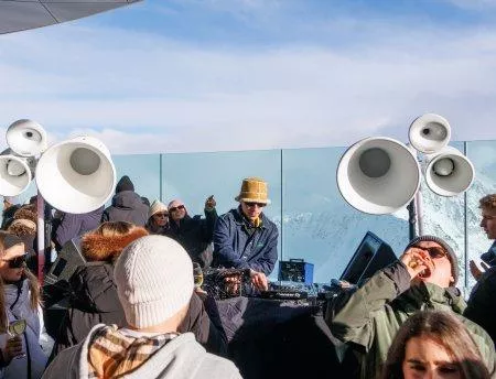 A lively outdoor party with a DJ and large speakers. The guests are enjoying the music and the sunny mountain scenery.