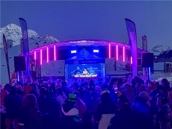 Eine große Menschenmenge feiert auf einem Berg bei einer Abendveranstaltung. Die Bühne ist mit bunten Lichtern beleuchtet und die schneebedeckten Berge sind im Hintergrund sichtbar.