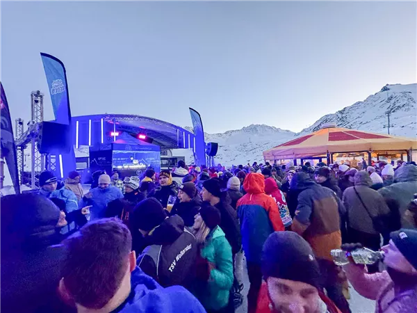 Eine lebhafte Menschenmenge vor einer Bühne in einer Berglandschaft. Im Hintergrund sind Schnee und Berge sichtbar, und es gibt Festbeleuchtung.