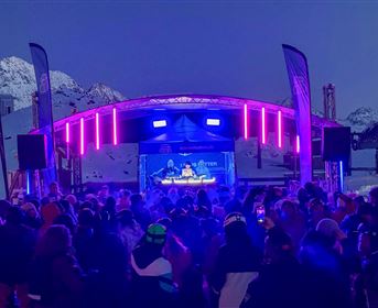 Eine große Menschenmenge feiert auf einem Berg bei einer Abendveranstaltung. Die Bühne ist mit bunten Lichtern beleuchtet und die schneebedeckten Berge sind im Hintergrund sichtbar.