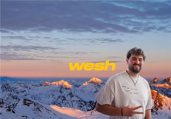 Ein lächelnder junger Mann steht vor einer beeindruckenden Berglandschaft mit schneebedeckten Gipfeln. Der Himmel hat eine sanfte Morgen- oder Abendfarbe.