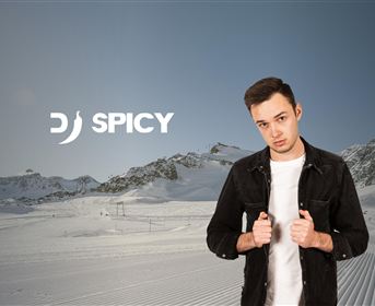Ein junger Mann steht in einer winterlichen Landschaft mit schneebedeckten Bergen im Hintergrund. Das Bild zeigt den Namen "DJ SPICY" in moderner Schrift.