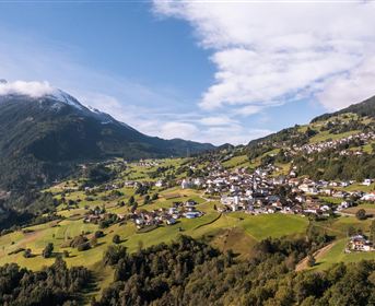 A picturesque mountain landscape with a small village in green valleys. In the background, snow-capped mountains and a clear sky can be seen.