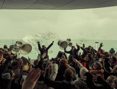 A lively party with a cheering crowd in the mountains. In the background, snow-capped peaks are visible.