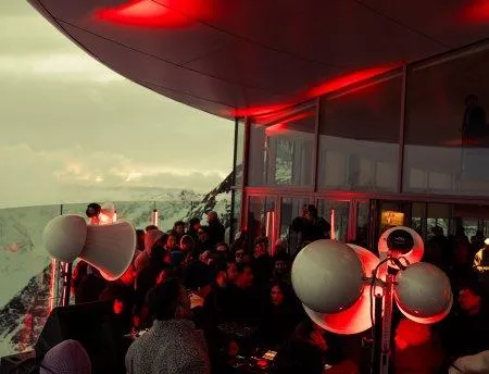 A lively event in a modern space with many people and red lights. In the background, mountains and clouds are visible.