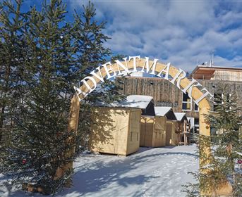 Ein Adventsmarkt mit einem großen Bogen und festlich dekorierten Holzständen. Umgeben von schneebedeckten Bäumen und einem klaren blauen Himmel.
