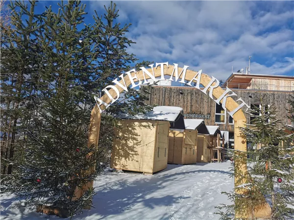 Ein Adventsmarkt mit einem großen Bogen und festlich dekorierten Holzständen. Umgeben von schneebedeckten Bäumen und einem klaren blauen Himmel.