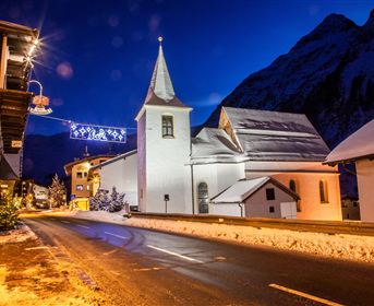 Eine malerische schneebedeckte Kirche bei Nacht. Die Umgebung ist festlich beleuchtet und von schneebedeckten Bergen umgeben.