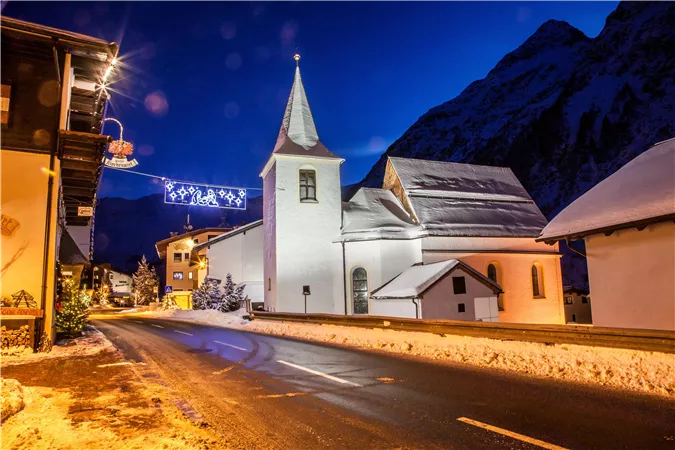 Eine malerische schneebedeckte Kirche bei Nacht. Die Umgebung ist festlich beleuchtet und von schneebedeckten Bergen umgeben.