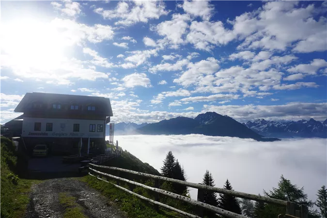 A house on a hill above a blanket of clouds overlooking the mountains. The sky is clear and blue with some white clouds.