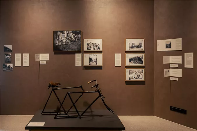 A modern museum with a sculptural bicycle installation in the foreground. Various pictures and informational texts can be seen on the walls.
