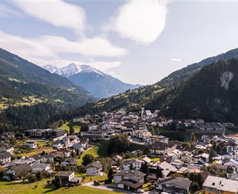 Eine malerische Berglandschaft mit einem kleinen Dorf im Tal. Umgeben von grünen Wiesen und hohen Bergen, darunter schneebedeckte Gipfel.