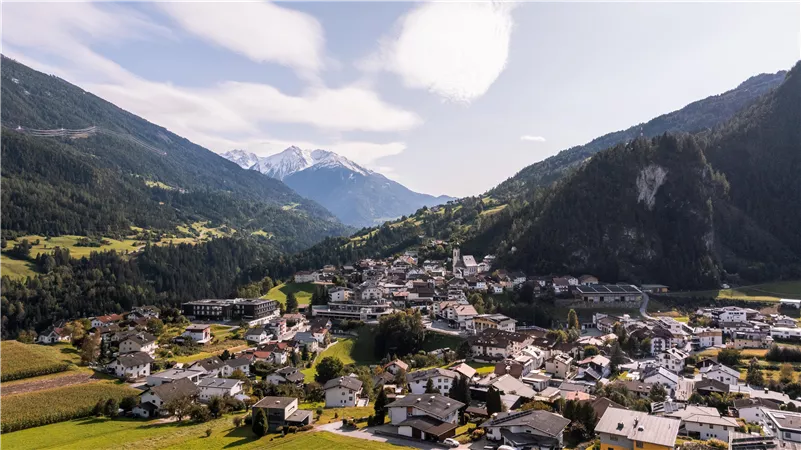 A picturesque view of a small village surrounded by mountains. The landscape is green and hilly with clear air and a blue sky.
