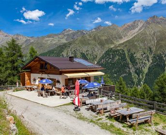 A cozy mountain hut nestled among green mountains. Outside, there are tables with sun umbrellas, and the surroundings are picturesque.