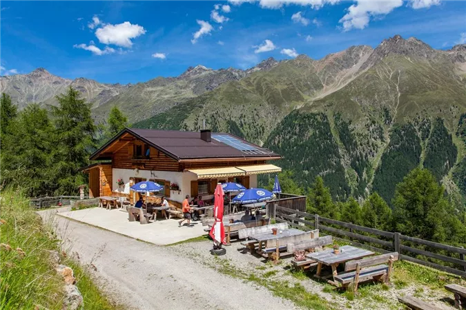 A cozy mountain hut nestled among green mountains. Outside, there are tables with sun umbrellas, and the surroundings are picturesque.