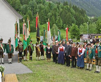 Eine Gruppe von Personen in traditioneller Kleidung versammelt sich im Freien. Sie halten Fahnen und stehen vor einer malerischen Berglandschaft.