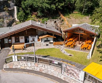 Eine rustikale Berghütte mit Holzveranda und Gartenmöbeln. Umgeben von Bäumen und Felsformationen.