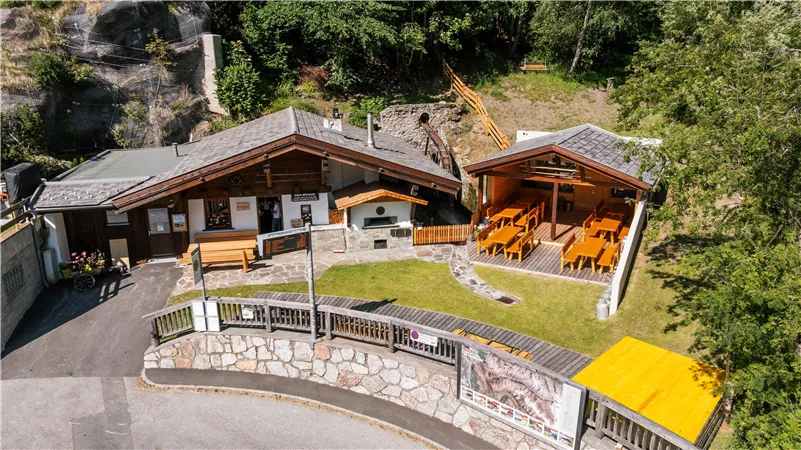 Eine rustikale Berghütte mit Holzveranda und Gartenmöbeln. Umgeben von Bäumen und Felsformationen.