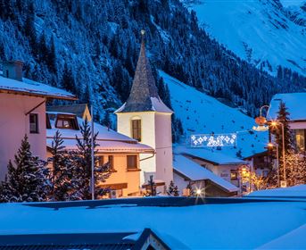 Eine malerische Winterlandschaft mit verschneiten Häusern und einem Kirchturm. Der Abendhimmel ist blau und die Lichter der Stadt strahlen in der Dämmerung.