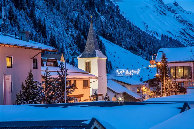 Eine malerische Winterlandschaft mit verschneiten Häusern und einem Kirchturm. Der Abendhimmel ist blau und die Lichter der Stadt strahlen in der Dämmerung.