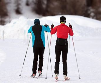 Two cross-country skiers are moving on a snow-covered track. In the background, snow-covered hills can be seen.