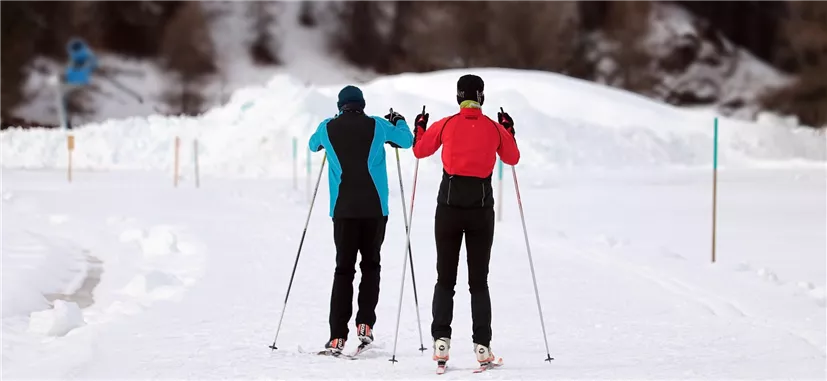 Two cross-country skiers are moving on a snow-covered track. In the background, snow-covered hills can be seen.