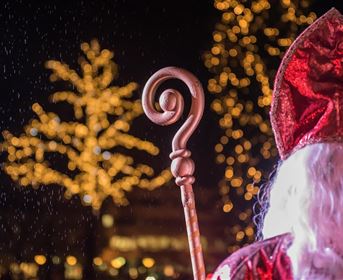 A mysterious man in a red cloak with a curved staff stands in the foreground. In the background, festive lights shine on a tree.