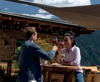 Ein Paar toastet mit Gläsern auf einer sonnigen Terrasse. Der Hintergrund zeigt eine schöne Berglandschaft.