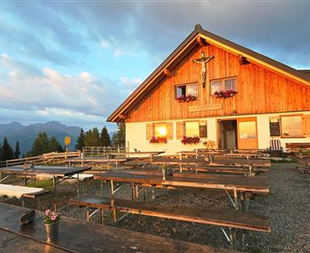 A rustic mountain house with a wooden facade and blooming flower boxes. In front of the house, several benches are placed on a gravel area, surrounded by a beautiful mountain landscape.