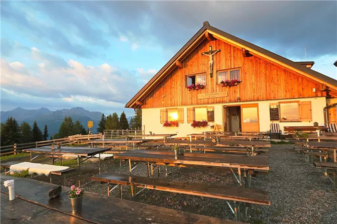Ein rustikales Berghaus mit Holzfassade und blühenden Blumenkästen. Vor dem Haus stehen mehrere Bänke auf einem Kiesplatz, umgeben von einer schönen Berglandschaft.