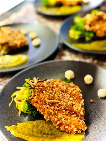 A deliciously arranged dish on a black plate. It features breaded fish with side dishes of vegetables and a sauce.