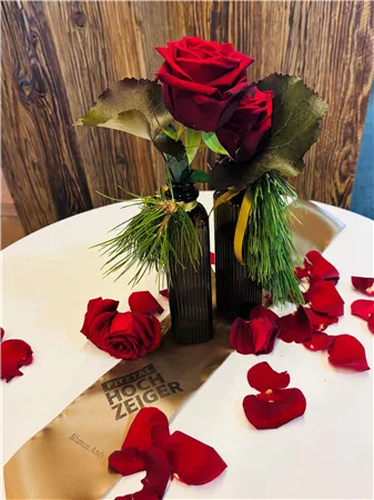 An elegant table with two roses in a decorative vase and scattered red rose petals. In the background lies a golden fabric with an inscription.