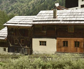Ein traditionelles Holzhaus in einer malerischen Berglandschaft. Es hat ein schützendes Dach und ist von grünen Wiesen und Bäumen umgeben.