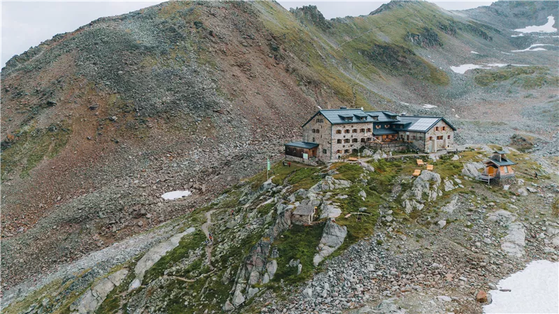 Ein Berghaus auf felsigem Gelände, umgeben von beeindruckenden Bergen. Die Landschaft ist rau und unberührt, mit wenigen Schneeflecken.