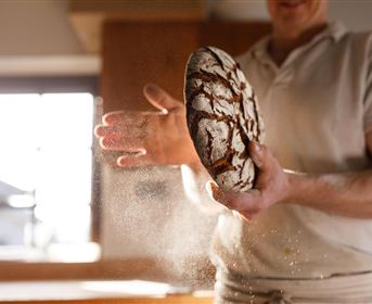 Ein Bäcker hält ein frisch gebackenes Brot in den Händen. Mehlstaub schwebt in der Luft.