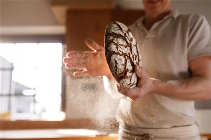 Ein Bäcker hält ein frisch gebackenes Brot in den Händen. Mehlstaub schwebt in der Luft.